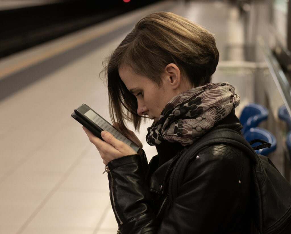 Photo de Laetitia assise dans une station de métro et lisant sur sa liseuse un livre. Laetitia porte une veste en cuir noir et une écharpe gris et mauve.