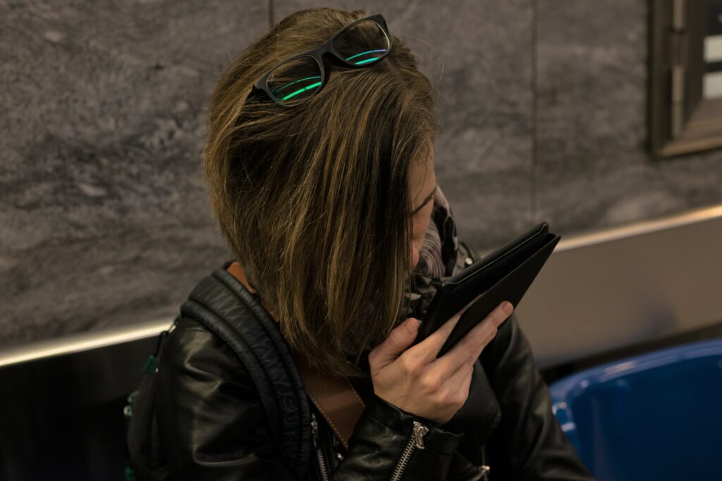 Photo de Laetitia assise dans une station de métro et lisant sur sa liseuse un livre. Laetitia porte une veste en cuir noir et une écharpe gris et mauve.